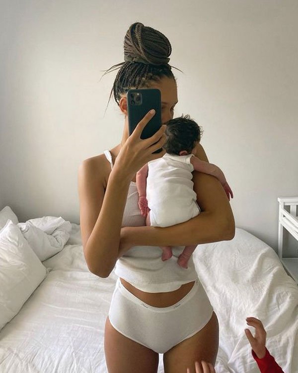 Mãe e bebê em quarto minimalista. Ela está usando roupa íntima branca e top de alças finas, que remetem a conforto e praticidade. O cabelo está com tranças estilizadas, um coque alto, evidenciando uma tendência de penteados protetivos e modernos.