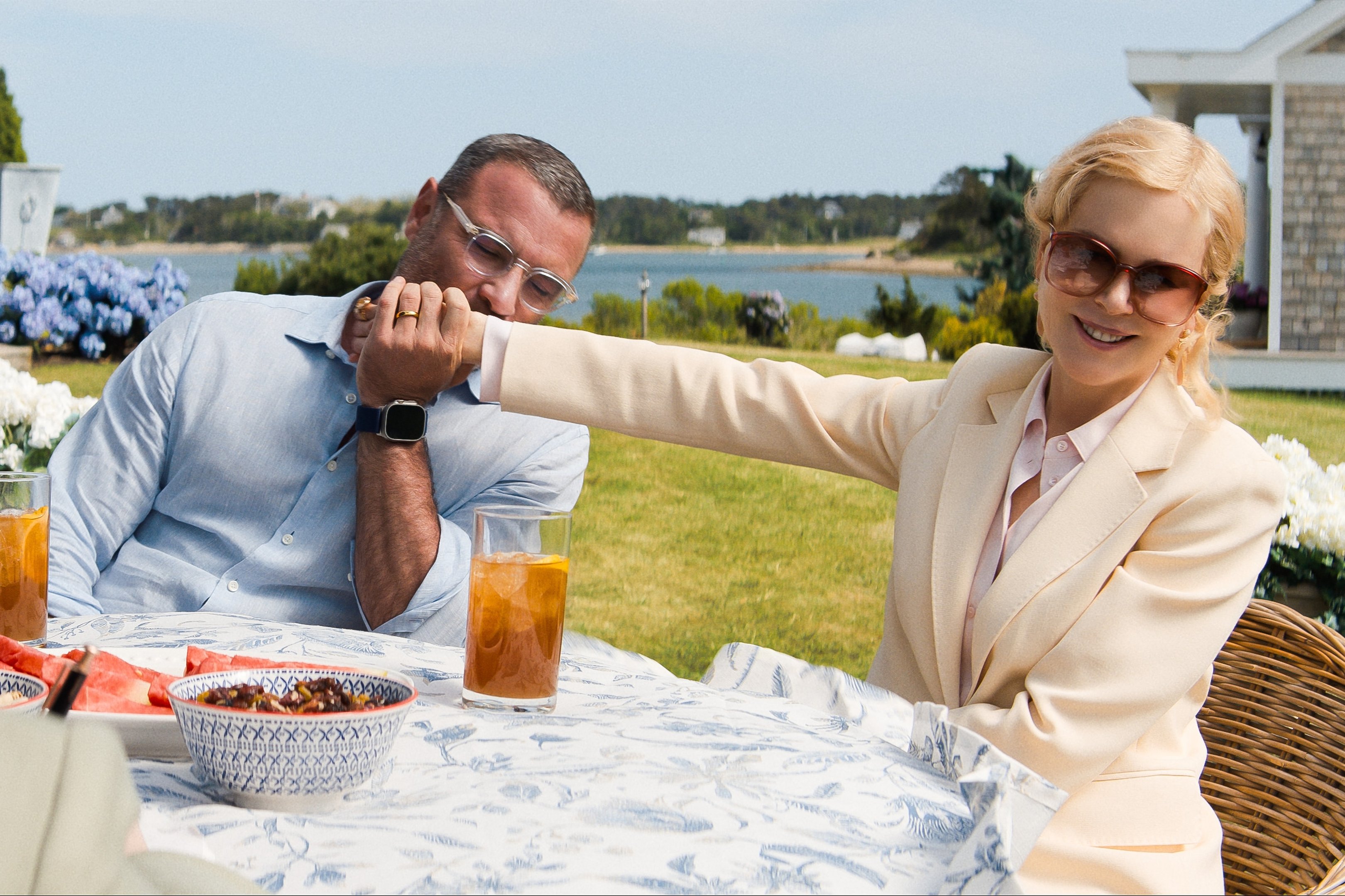 Imagem de casal ao ar livre. Homem usa camisa social azul clara e óculos de grau; mulher veste um elegante blazer nude, camisa rosa clara e óculos de sol grandes.