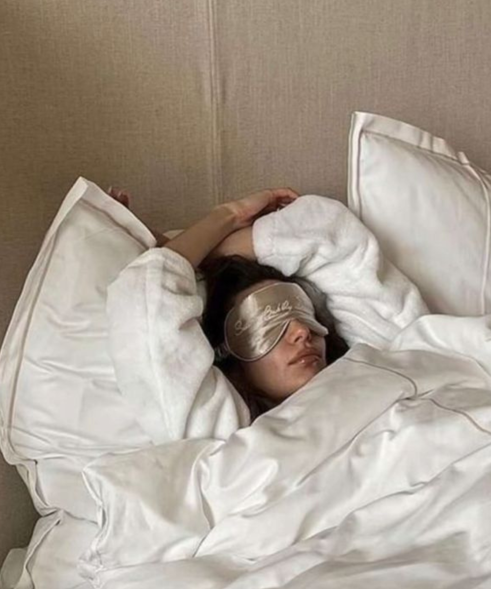 Pessoa deitada com uma máscara de dormir de cetim e roupão branco de tecido macio, em uma cama com lençóis, travesseiros e edredom brancos. O estilo transmite conforto e tendência hygge de bem-estar e relaxamento.