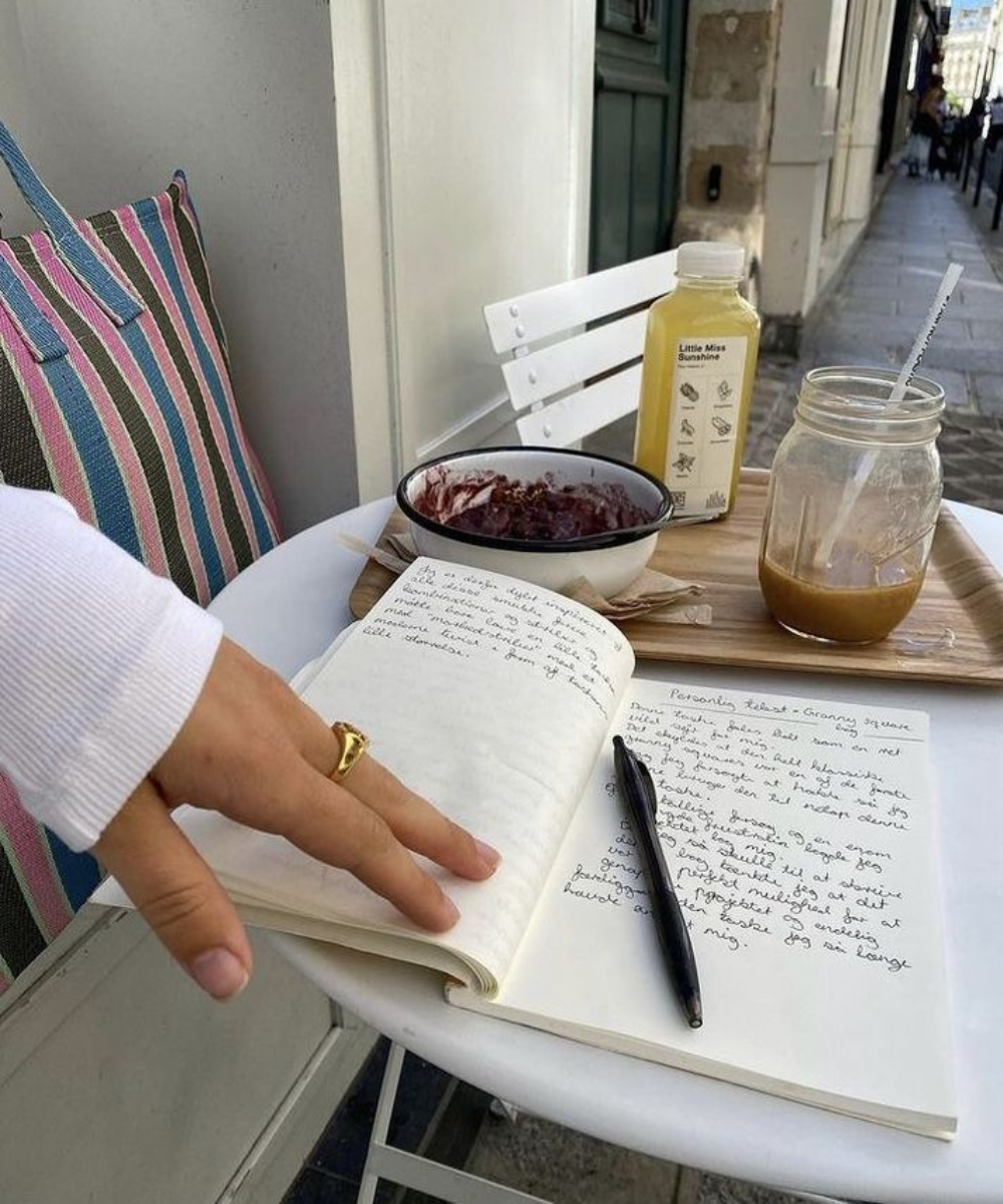 Imagem mostrando uma cena de café ao ar livre. Tendências de moda: uso de anel dourado discreto, caderno para anotação, bebidas em frascos reutilizáveis e bolsa listrada grande. O ambiente sugere um estilo casual e consciente, focado na sustentabilid