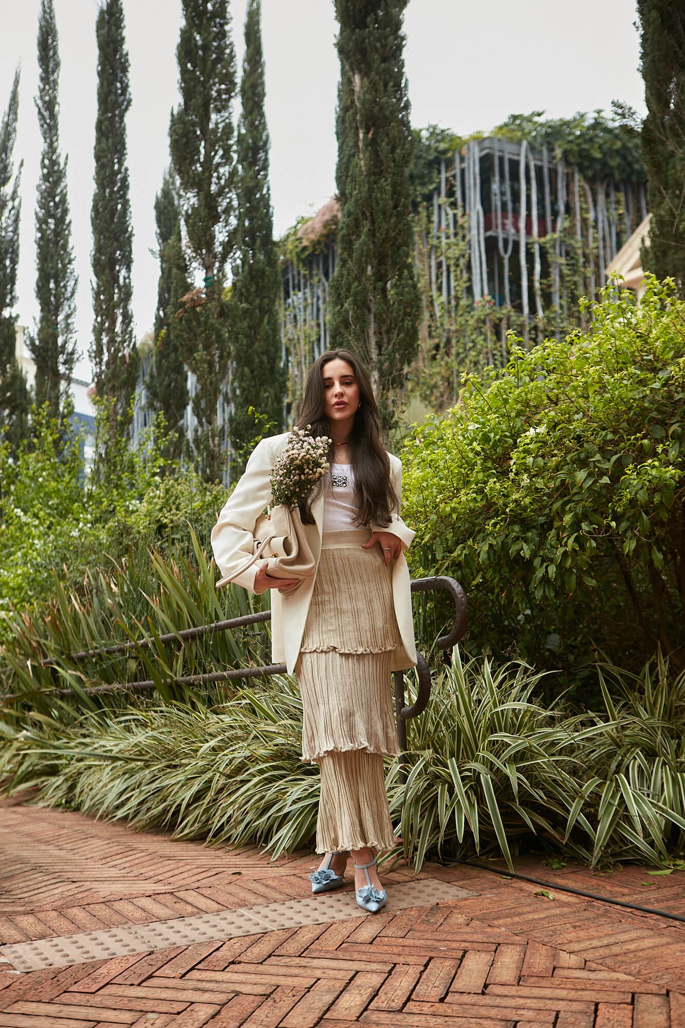 Jovem usando blazer branco elegante, top branco, saia plissada em tons neutros e sapatos de salto azul claro. Mistura de estilos clássico e moderno com toques românticos. Perfeito para ocasiões formais ao ar livre.