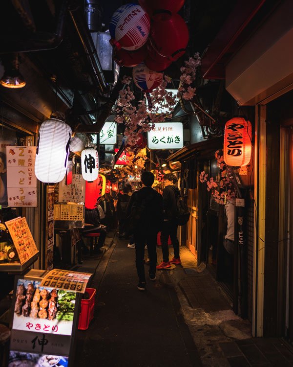 Imagem de um beco iluminado com lanternas coloridas. Pessoas vestem roupas casuais, como jaquetas e calças escuras. Predominância de negros e tonalidades escuras sugerem moda urbana, contrastando com o ambiente vibrante.