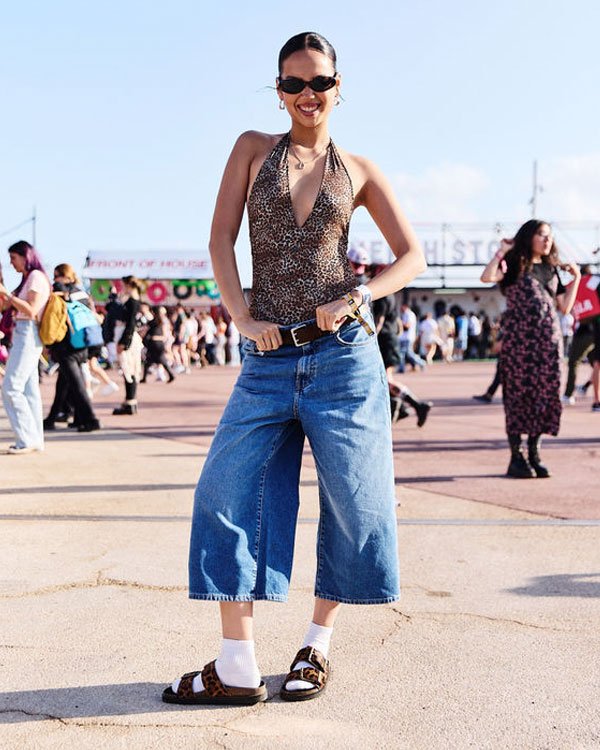 Look ousado e moderno com tendências de moda urbana. A mulher veste uma blusa com estampa animal print e calça jeans oversized de cintura alta e boca larga. O look é complementado por sandálias com meia branca e óculos de sol estilizados.