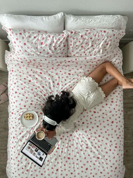 Pessoa usando pijama de renda branca, deitada em cama com lençóis e fronhas estampados com pequenos corações. A cena inclui notebook, bebida quente e fones de ouvido brancos, retratando conforto e estilo caseiro moderno.