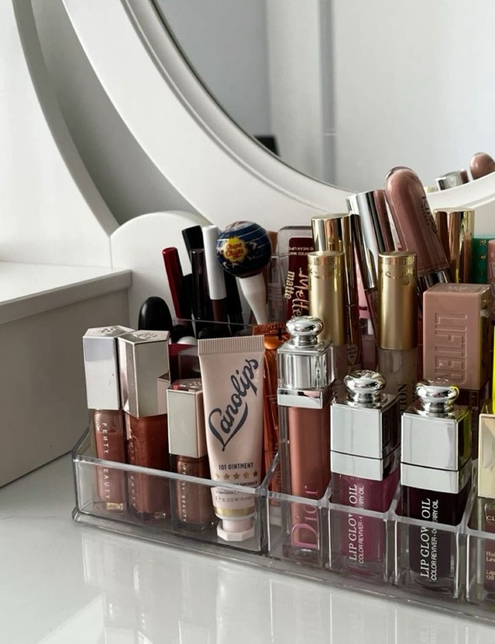 Organizador de maquiagem com diversos produtos de beleza, incluindo batons, lip gloss e hidratantes labiais de marcas populares. Tendência de moda em maquiagens e cuidados com os lábios evidenciando tons nude e embalagens elegantes.