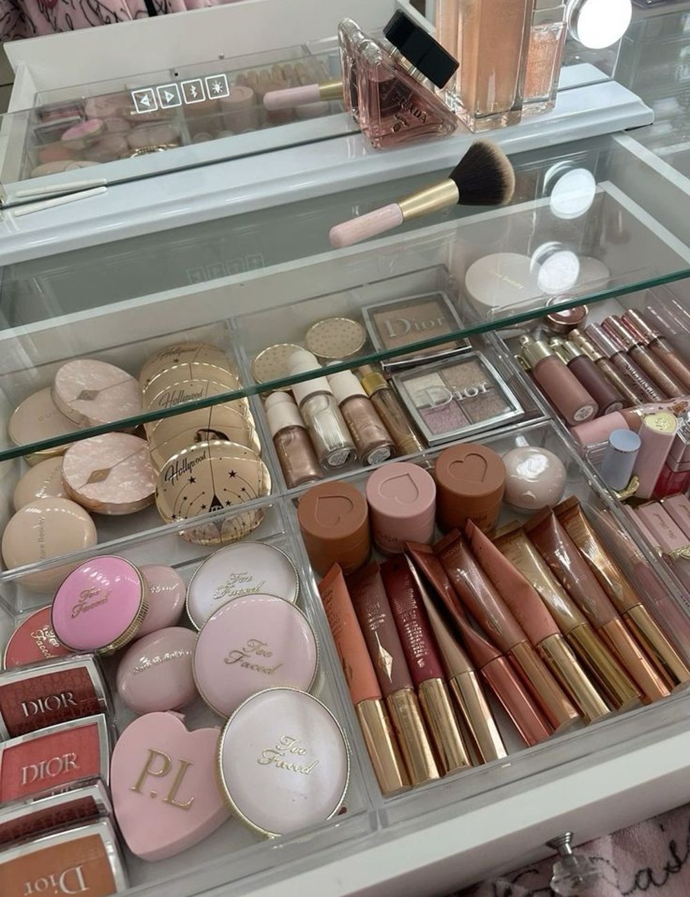 Organizador de maquiagem com diversos produtos de maquiagem de marcas renomadas como Dior e Too Faced. Itens incluem pós compactos, blushes, batons, glosses, bases e pincéis em tons nude, rosa e dourado.