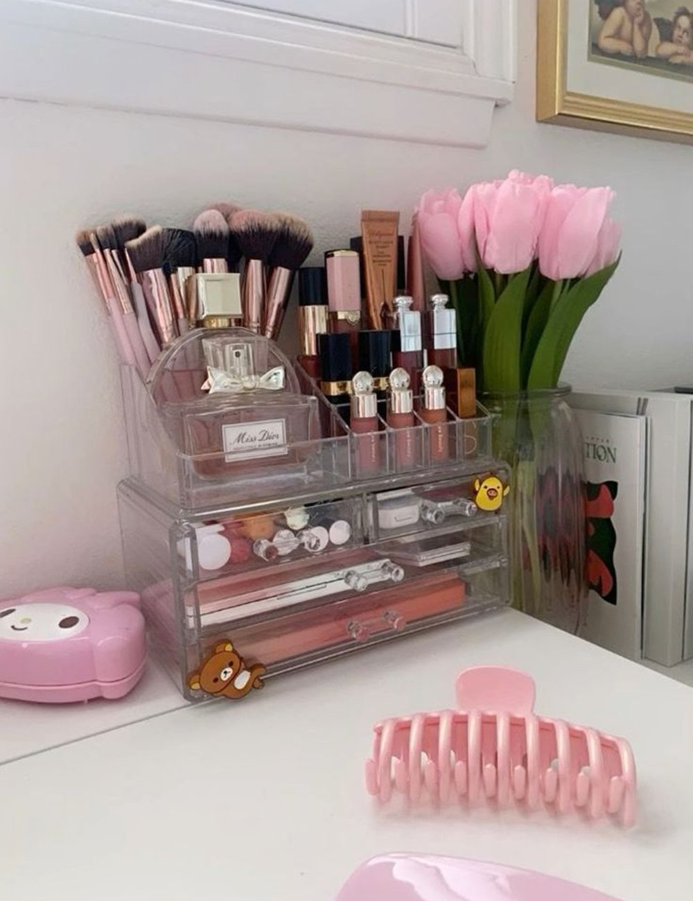 Organizador de maquiagem transparente com pincéis, batons, perfumes e produtos de beleza, predominando tons de rosa e dourado. Decorado com adesivos fofos e tulipas rosas, reflete tendências de estética clean e kawaii.