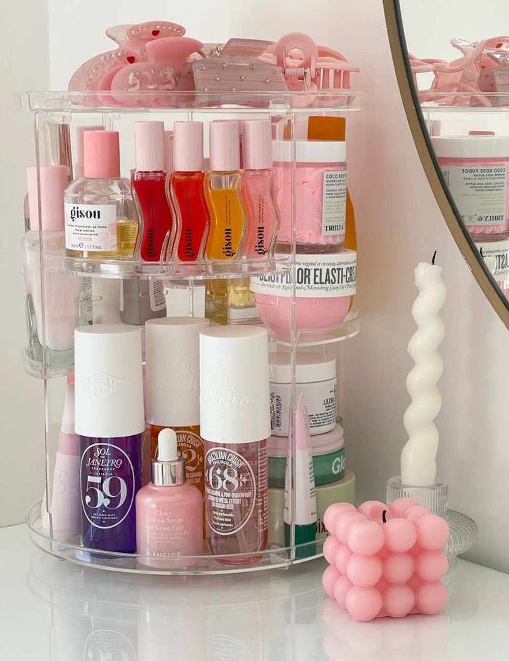 Organizador de maquiagem com produtos de beleza em tons de rosa, roxo e branco. Destaque para velas decorativas modernas e prendedores de cabelo. Itens remetem a uma estética clean e feminina, com tendência minimalista e contemporânea.