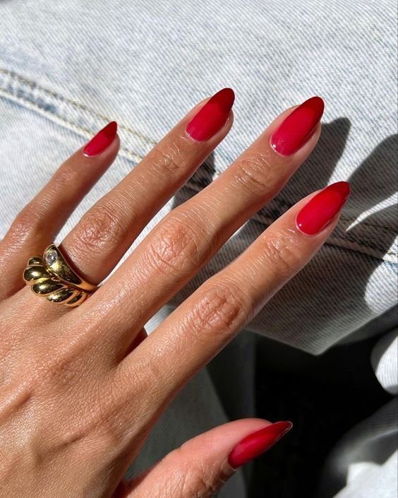 Unhas com esmalte vermelho ombré, tendência moderna em nail art, combinadas com anel dourado robusto. O fundo de jeans claro contrapõe o visual sofisticado das unhas, destacando um estilo casual-chic.