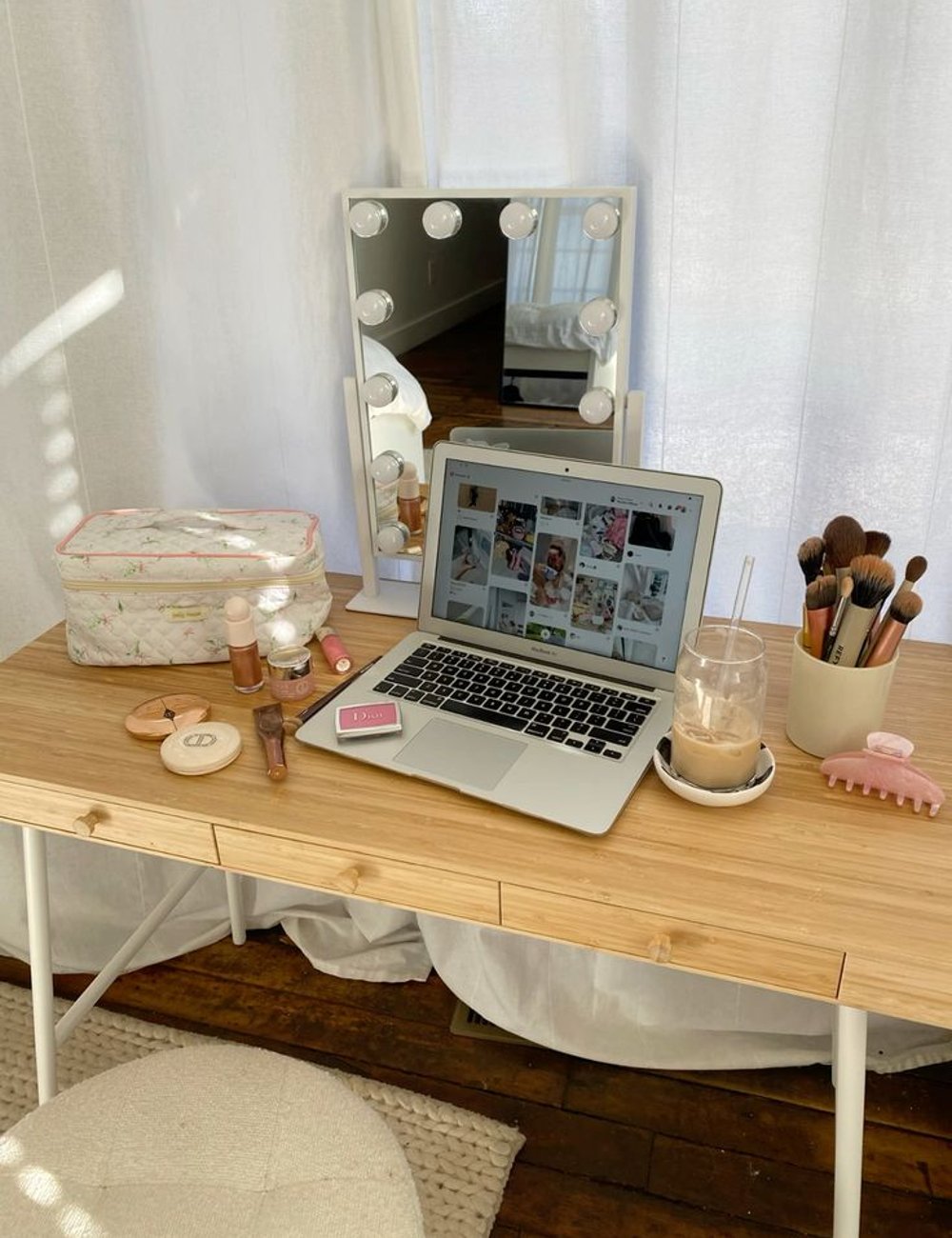 Imagem de penteadeira minimalista com espelho iluminado, kit de pincéis de maquiagem em xícara, laptop com página de moda aberta, cosméticos sobre a mesa e copo de café gelado. Decoração clean e organização de maquiagem.