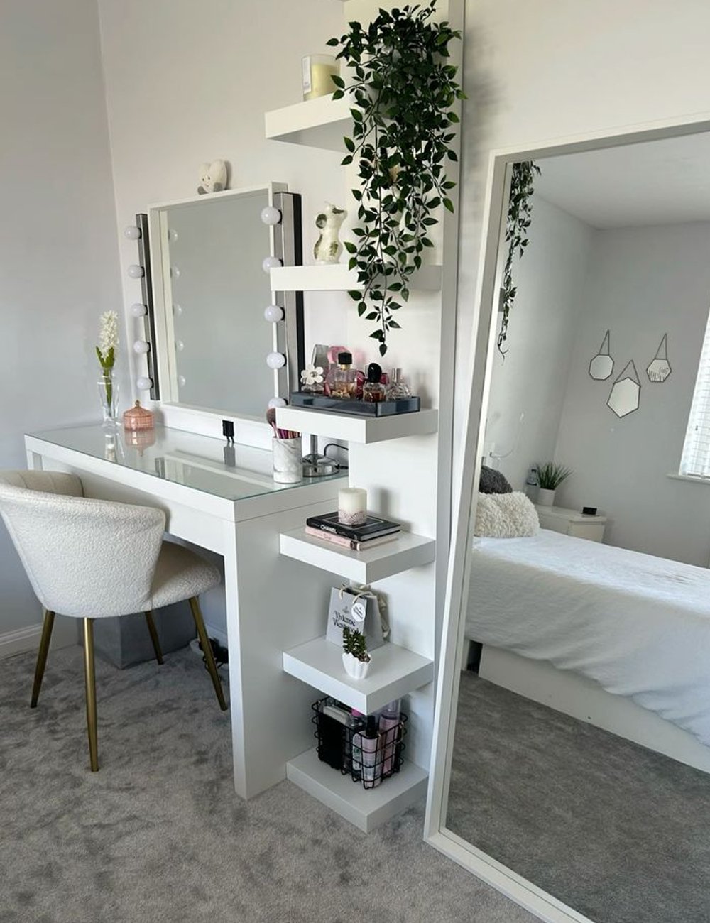 Quarto moderno com penteadeira branca minimalista decorada com plantas, espelho grande com luzes de camarim. Estilo clean com cadeira de veludo e pés dourados. Prateleiras organizadas com maquiagem, livros e velas. Ambiência elegante e aconchegante.