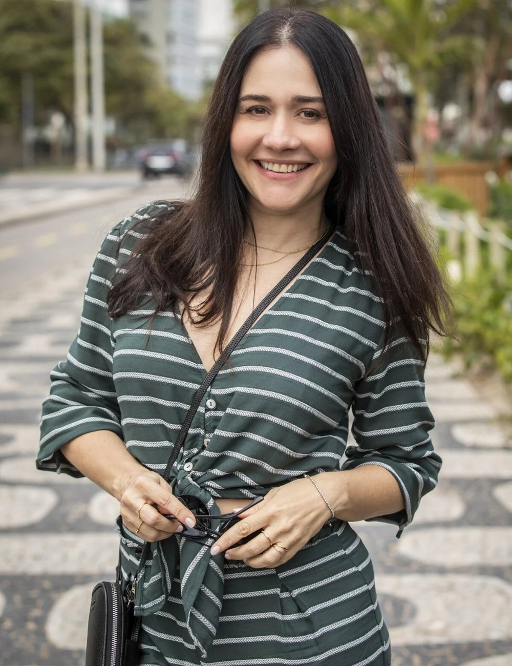 Alessandra Negrini vestindo uma blusa listrada com mangas 3/4 e amarração na cintura, em tons de verde e branco. A combinação casual e chic é complementada por acessórios discretos, como pulseiras e um colar delicado.