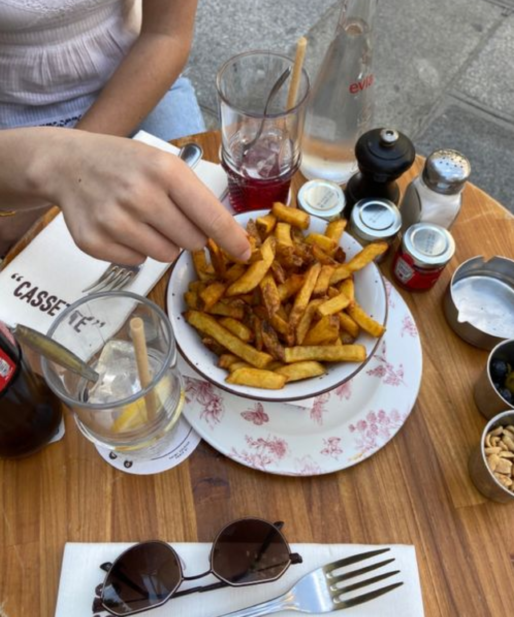 Pessoa vestindo blusa branca e jeans comendo batatas fritas em um ambiente casual. Na mesa há óculos de sol, garrafas de bebida e acessórios de mesa. Look descontraído e moderno, ideal para dias de verão em bistrôs ou cafés ao ar livre.