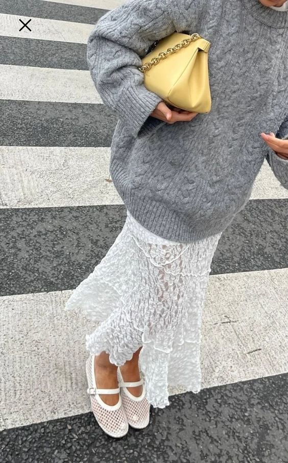 Look casual com suéter cinza oversized de tricô, saia midi branca com textura rendada e sapatos brancos com tiras e detalhes vazados. Acessório inclui bolsa amarela de correntes douradas.