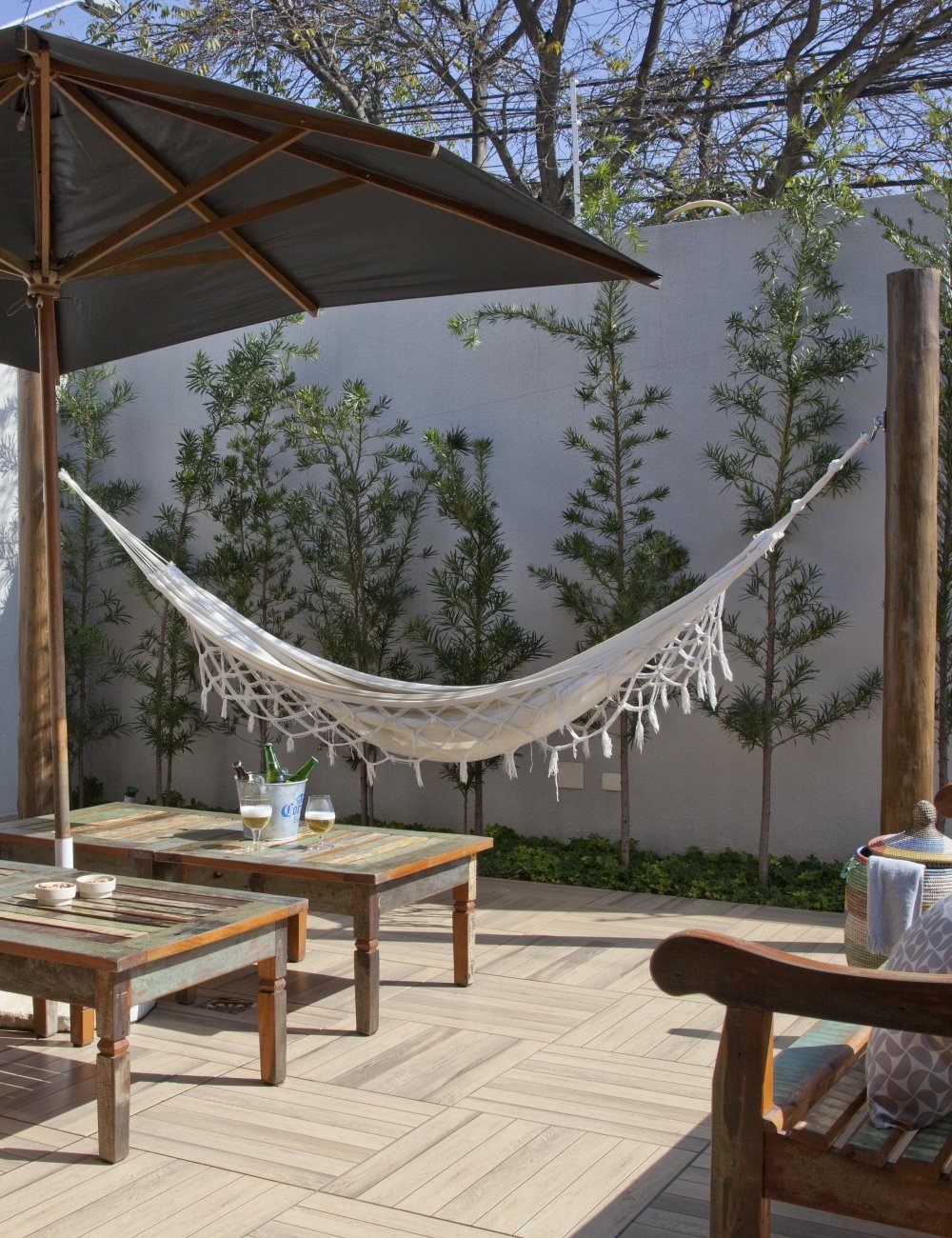 Área externa com rede branca de crochê, mesas rústicas de madeira e guarda-sol. Tendências de decoração incluem uso de móveis artesanais e estilo boho. Ambiente relaxante ao ar livre com elementos naturais e confortáveis. Piso ideal.