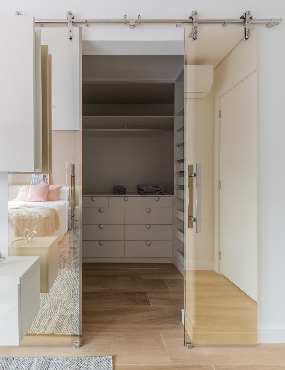 Guarda-roupa planejado com portas deslizantes de vidro transparente. O closet é moderno e minimalista, combinando tons neutros e madeira clara. Roupa de cama em tons suaves adiciona elegância ao ambiente. Piso ideal.