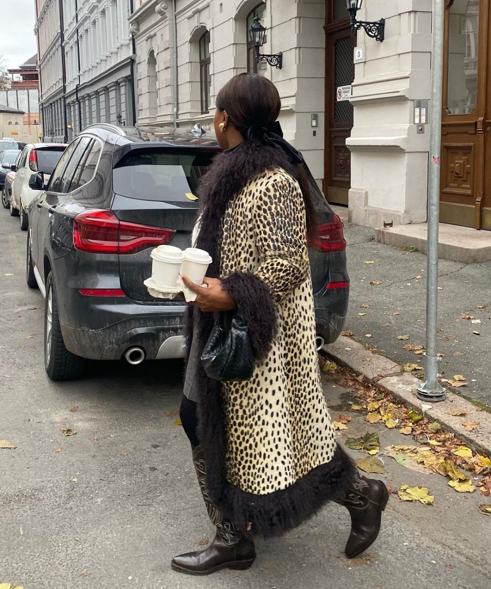 Pessoa usando casaco de pele com estampa de leopardo e detalhes de pele preta, combinando com botas marrons de couro e bolsa preta. Look elegante de inverno, destacando tendências de moda com estampas de animais e acessórios clássicos.