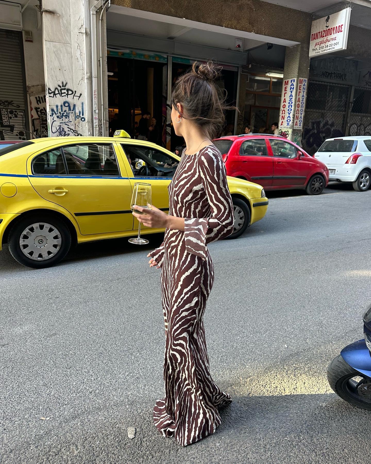 Mulher usando vestido longo com estampa animal print, tendência marcante em 2023. A peça, elegante e ousada, é combinada com cabelo preso em um coque alto, destacando um visual sofisticado e fashionista. Bebendo vinho em uma taça, em ambiente urbano.