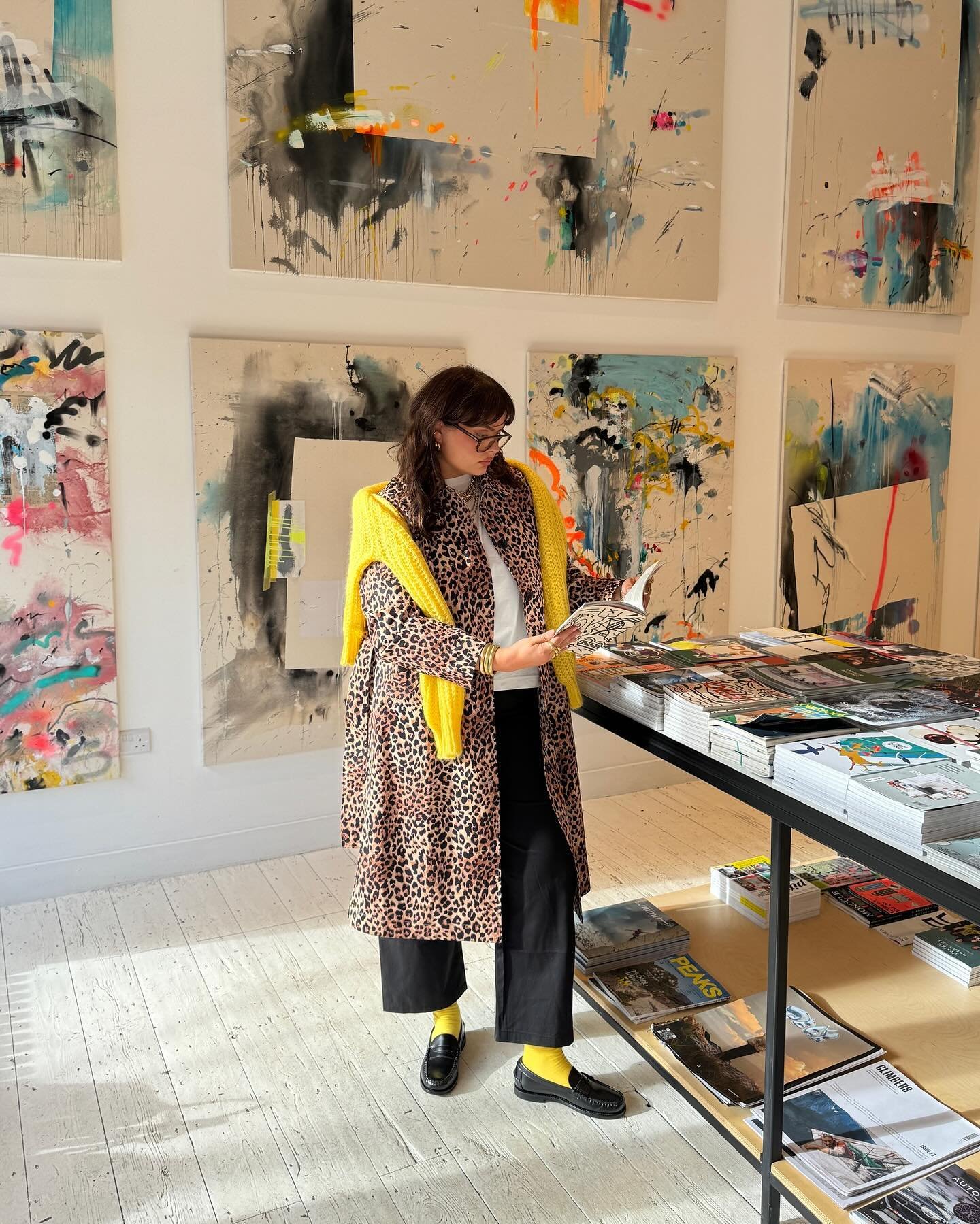 Pessoa em galeria de arte veste casaco de estampa animal print, suéter amarelo, calça preta e meia amarela. Combina tendências ousadas, misturando padrões e cores. Visual moderno com sapatos pretos clássicos e óculos de grau.