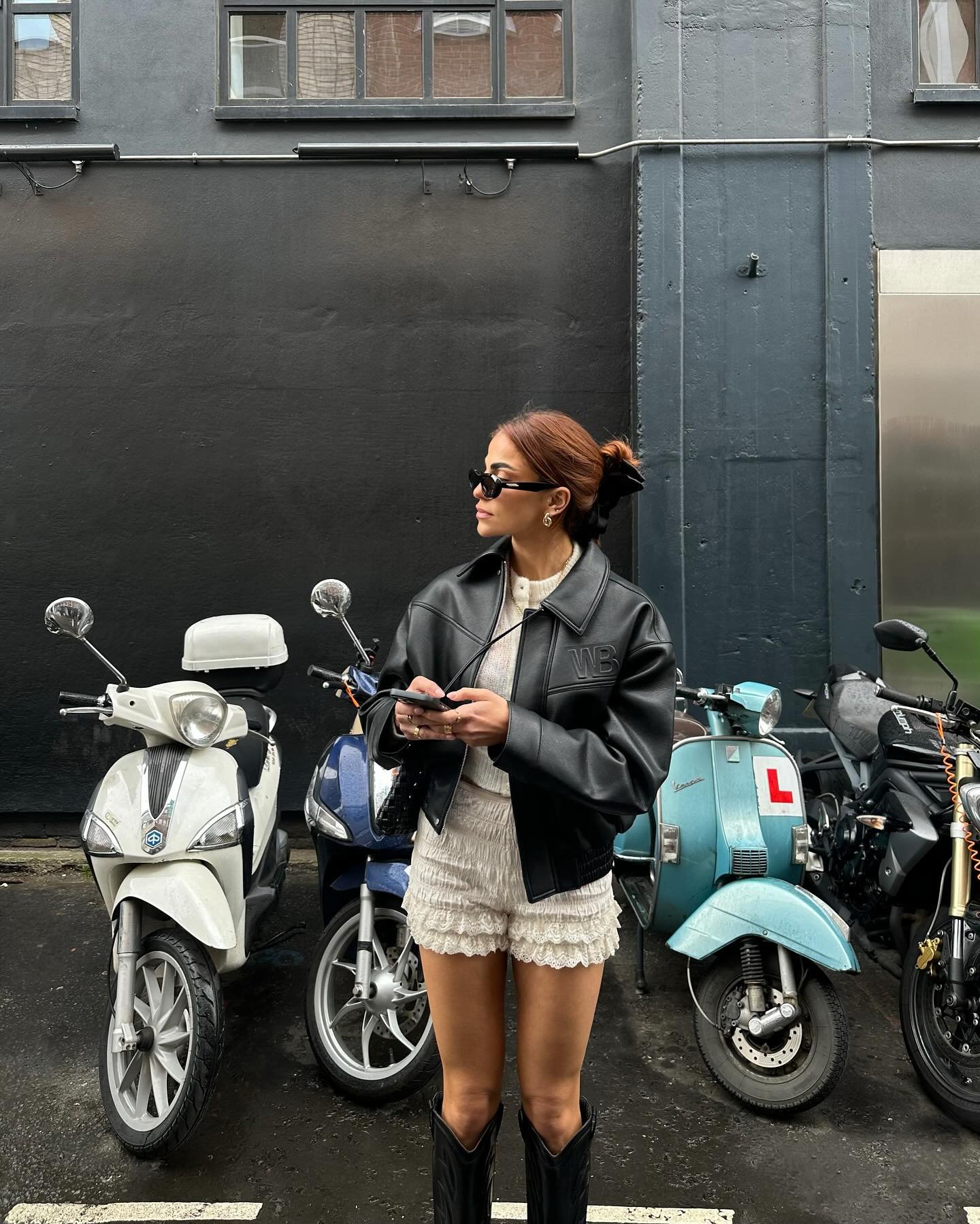 Pessoa posa na frente de motocicletas usando jaqueta de couro preta oversized, shorts de renda branco, óculos de sol e botas pretas de cano médio. Estilo urbano moderno, combinando peças retrô com tendências atuais.