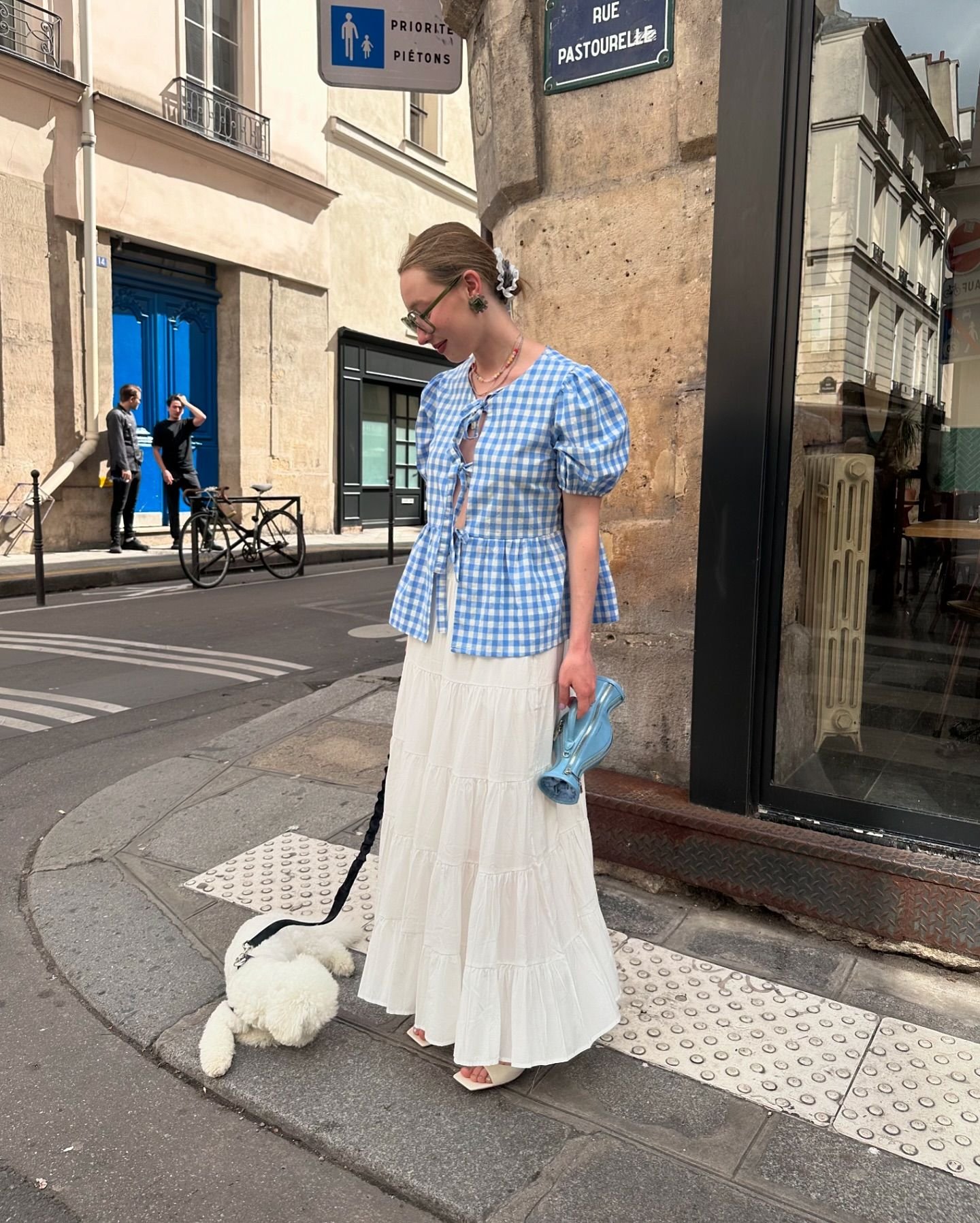 Mulher vestindo uma blusa xadrez azul e branca com mangas bufantes e amarração frontal, combinada com uma saia longa e branca. Ela carrega uma bolsa azul e usa óculos de sol modernos, enquanto passeia com um cachorro. Estilo casual e romântico.