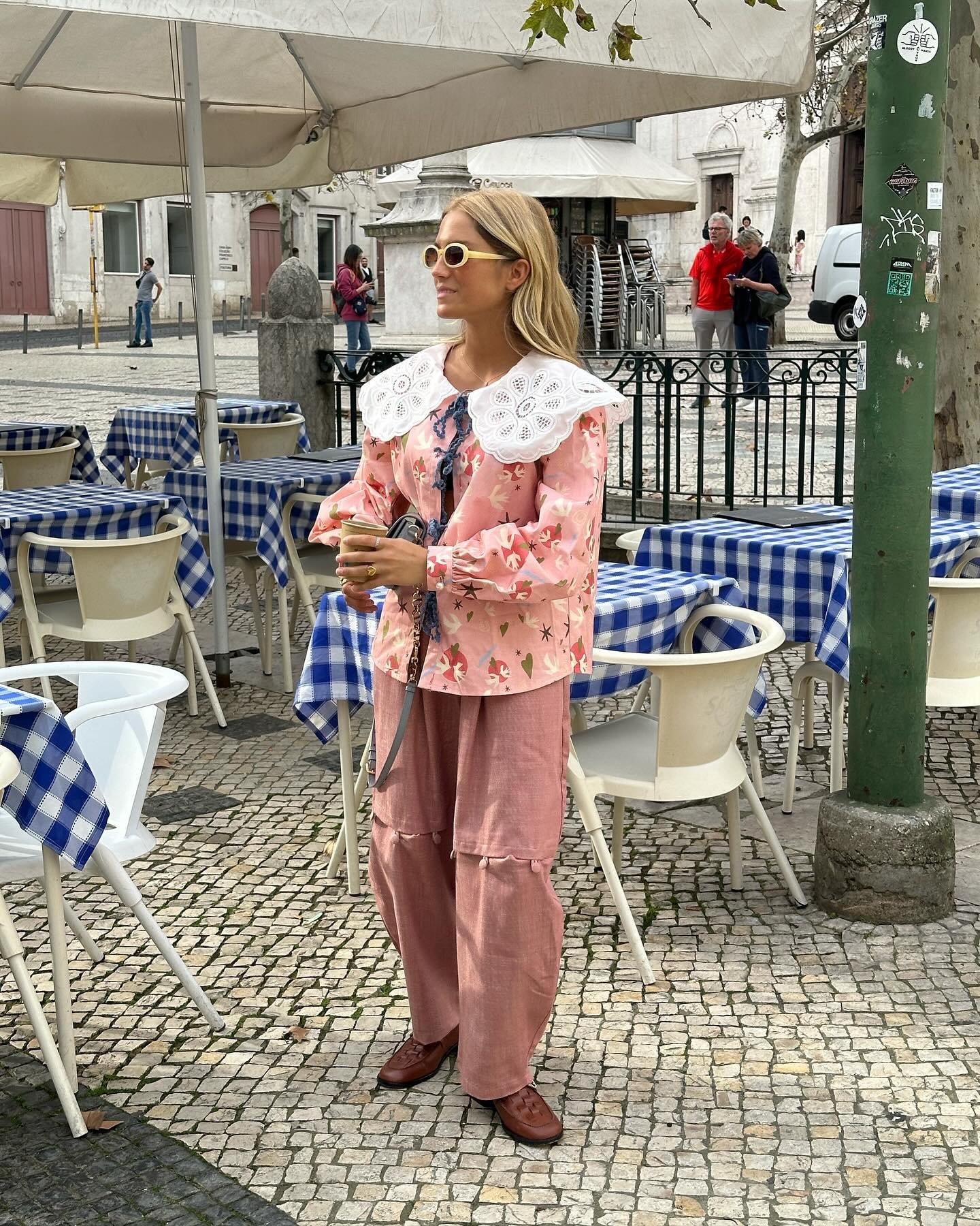 Mulher com blusa rosa floral de babados e gola rendada, calça larga rosa com bolsos, óculos de sol branco vintage e sapatos marrons. Estilo boêmio e romântico, com toque retrô. Ambiente de café ao ar livre.