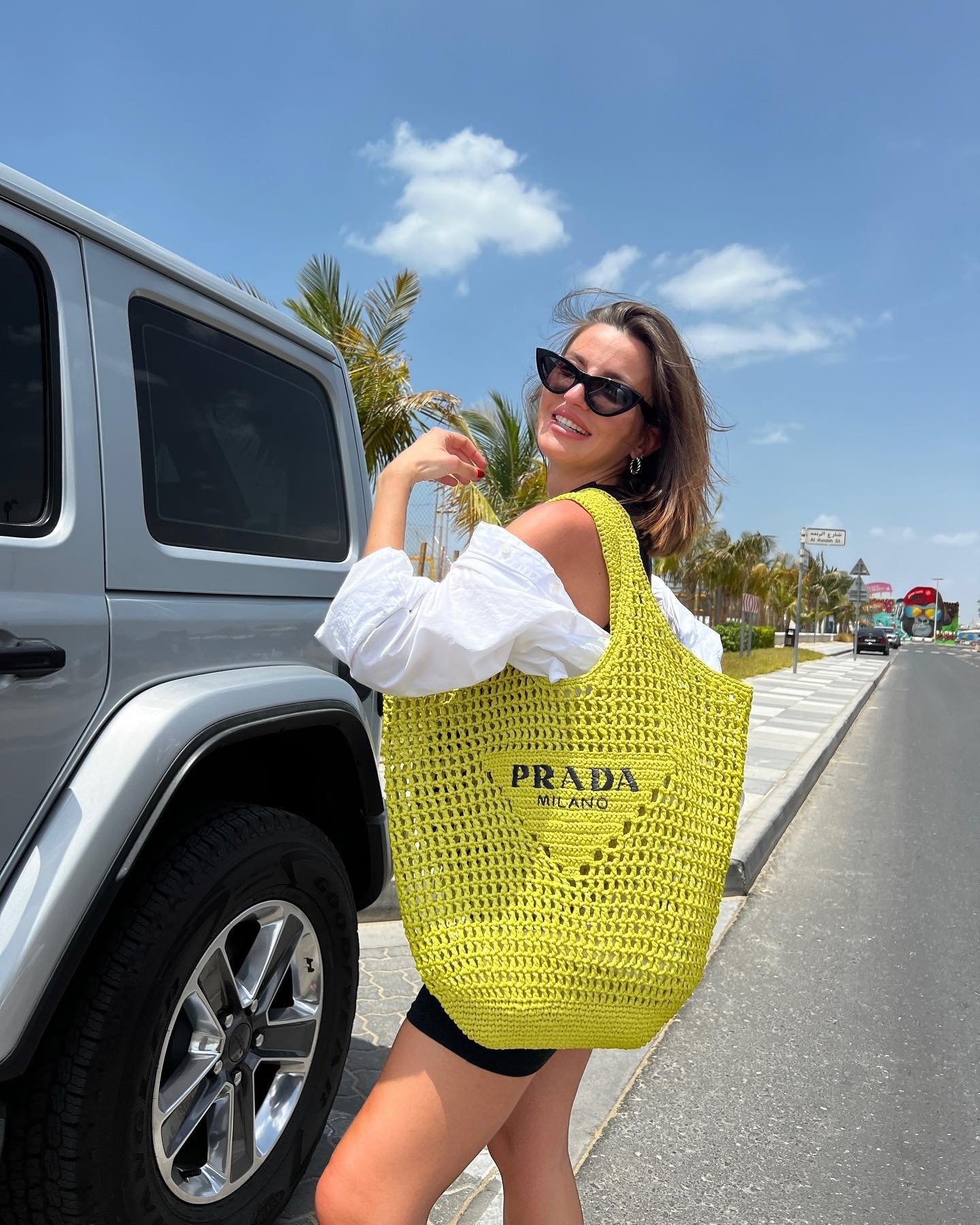 Mulher usa blusa branca ombro caído, shorts preto e bolsa de crochê amarela da Prada. Completa o look com óculos de sol modelo 