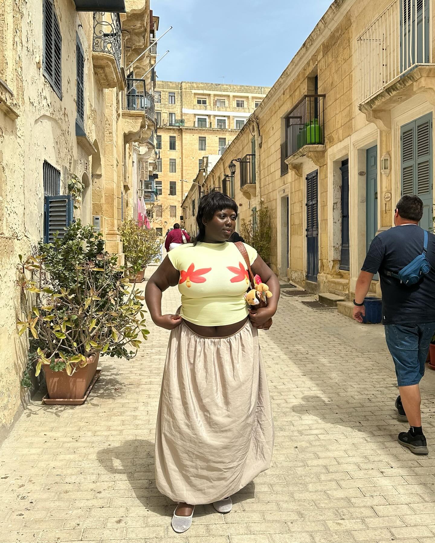 Mulher em rua histórica, veste top cropped com estampa de margaridas e saia longa bege, combinados com sapatilhas brancas. Look casual e descontraído, ideal para clima quente. Estilo mistura conforto e tendência floral.