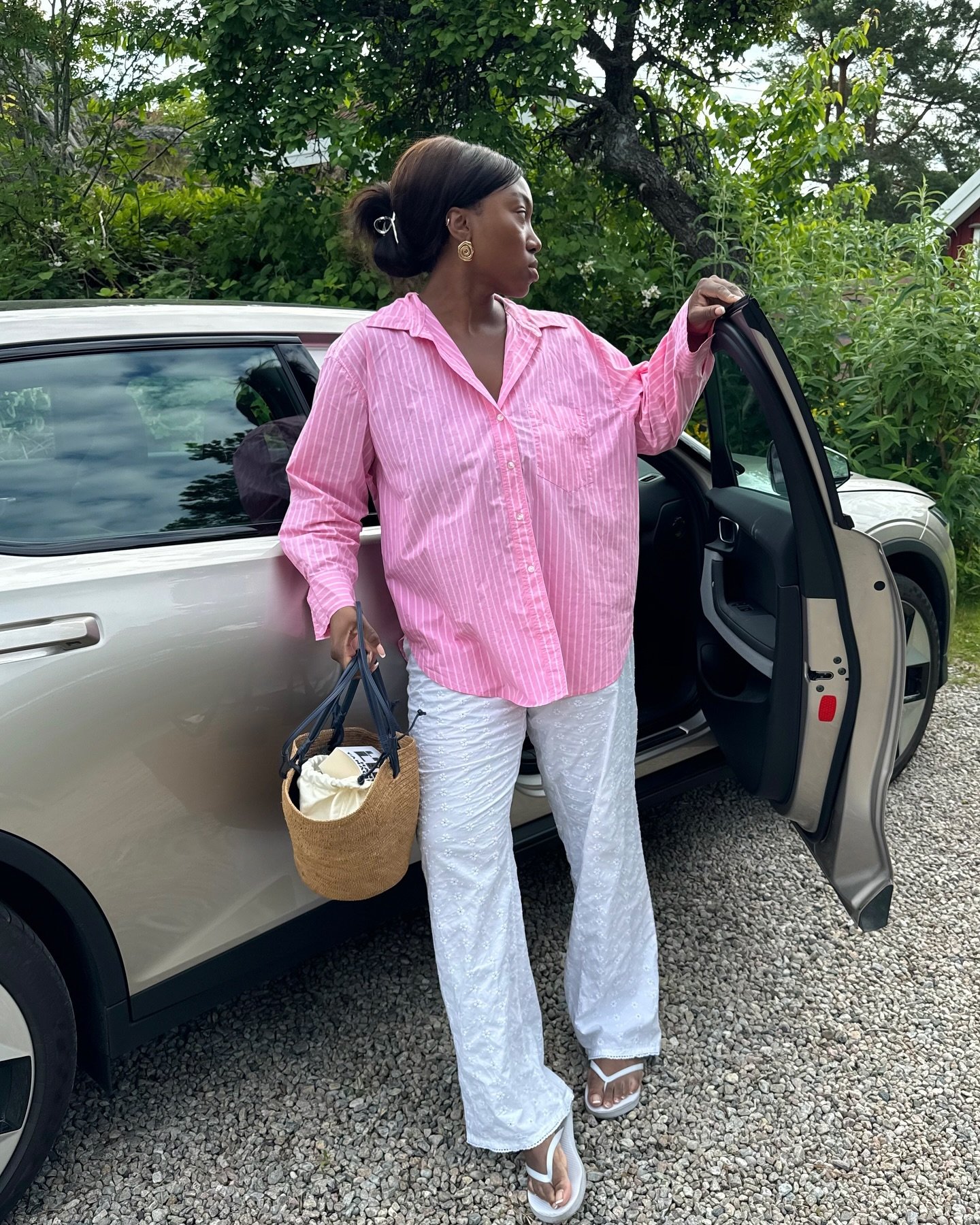 Pessoa usando uma camisa oversize rosa com listras brancas, calça branca texturizada e chinelos brancos. Ela complementa o look com uma bolsa de palha e brincos dourados, combinando conforto e elegância casual. Estilo descontraído.