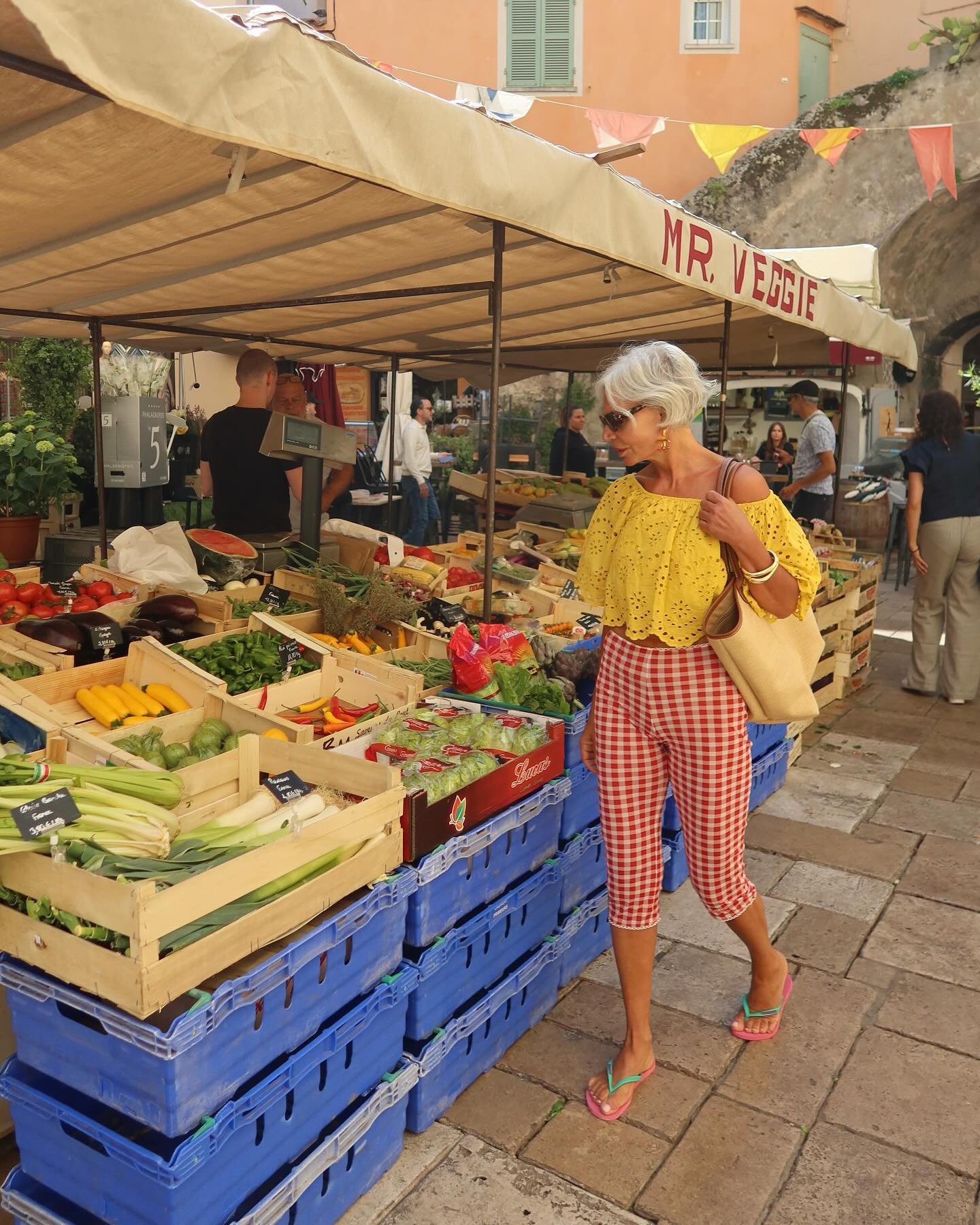 Mulher em feira de legumes usando look casual e colorido: blusa amarela rendada com ombros de fora, calça xadrez vermelha e branca estilo capri, chinelos de dedo multicoloridos e bolsa grande de palha. Estilo descontraído e alegre, ideal para passeios diurnos.