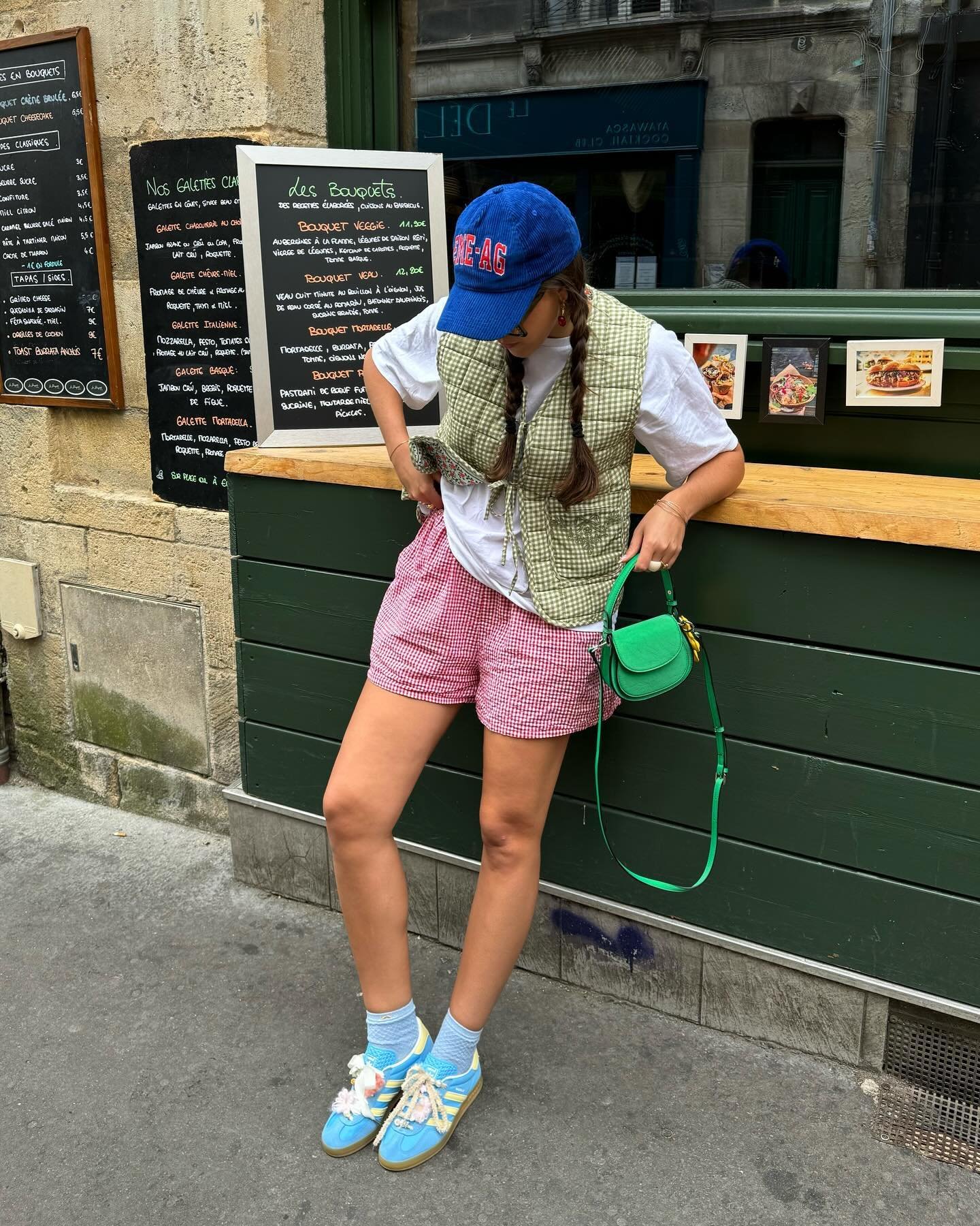 Jovem com estilo casual e despojado. Usa boné azul, camiseta branca, colete xadrez verde, shorts vermelhos de vichy, tênis coloridos com meias azuis e bolsa verde. Look mistura peças vintage e modernas. Ideal para indexação de moda urbana e tendências contemporâneas.