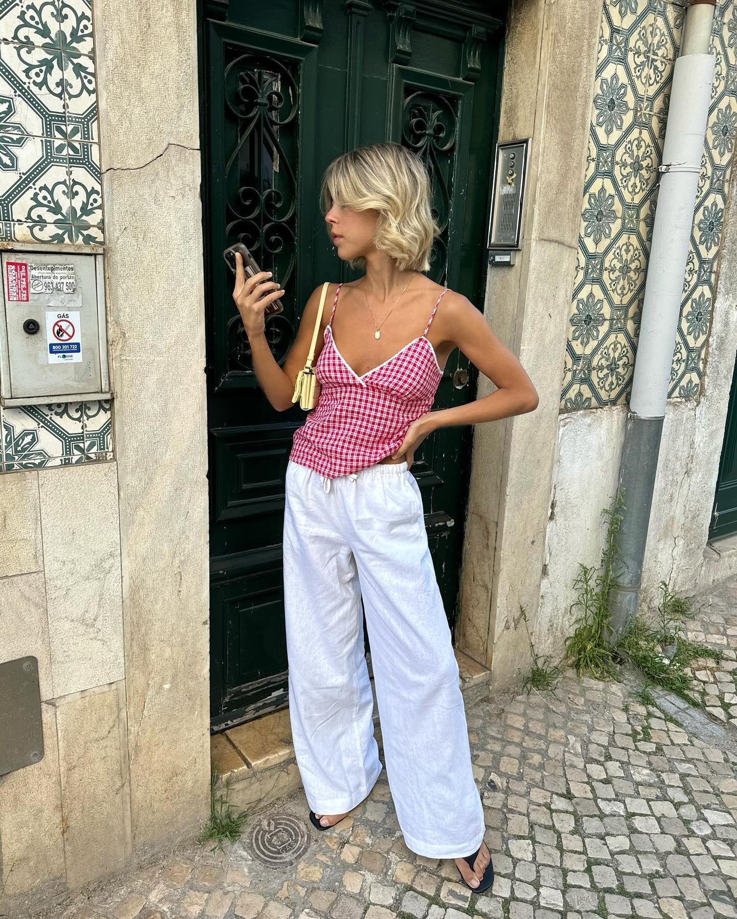 Pessoa com top xadrez vermelho e branco de alças finas, calça branca larga de tecido leve e sandálias pretas. Bolsa amarela pequena e cabelo loiro cacheado curto. Look casual e verão, ideal para passeios em dias quentes, combinando conforto e estilo.