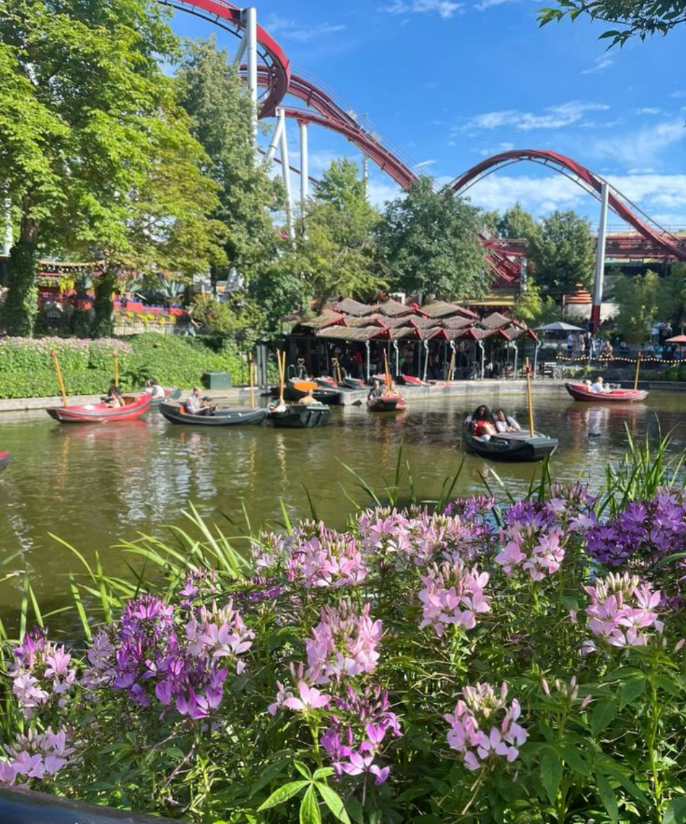 Parque de diversões com lago e pedalinho. Visitantes usam roupas leves e confortáveis, como camisetas e shorts, e seguem tendência de moda casual e prática para dias de verão. Colorido do parque contrasta com a natureza ao redor. Flores complementam