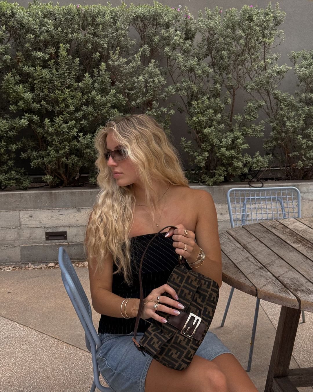 vestindo top preto tomara que caia, saia jeans e acessórios dourados. Bolsa de ombro com estampa monogramada e óculos de sol estilo retrô. Cabelo longos e ondulados. Estilo casual chic com toque vintage e moderno. queda por quebra