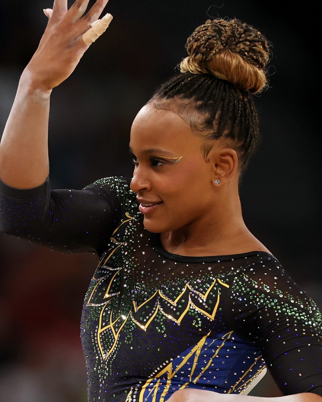 Atleta com penteado afro em coque alto e maquiagem elaborada. Veste collant de ginástica preto com detalhes brilhantes em verde, azul e dourado, enfatizando a elegância e o glamour das competições de ginástica rítmica. olimpíadas 2024