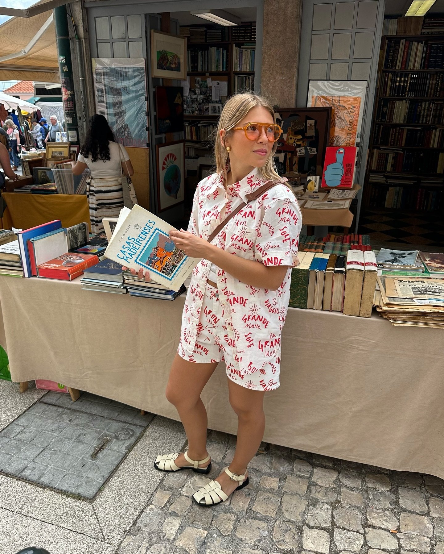 Jovem em um mercado de livros usados usa conjuntinho de camisa e short com estampa de palavras em vermelho, sandálias brancas de tiras cruzadas e óculos de sol com lentes alaranjadas.