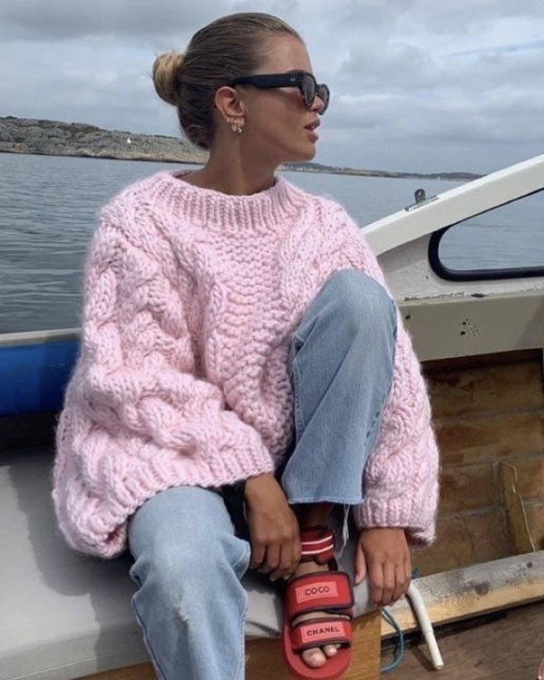 Mulher usando suéter rosa de tricô grosso, combinando com jeans claros e sandálias vermelhas Coco Chanel. Óculos de sol escuros e penteado coque. Estilo casual e confortável para dias frios, tendência de moda outono/inverno.