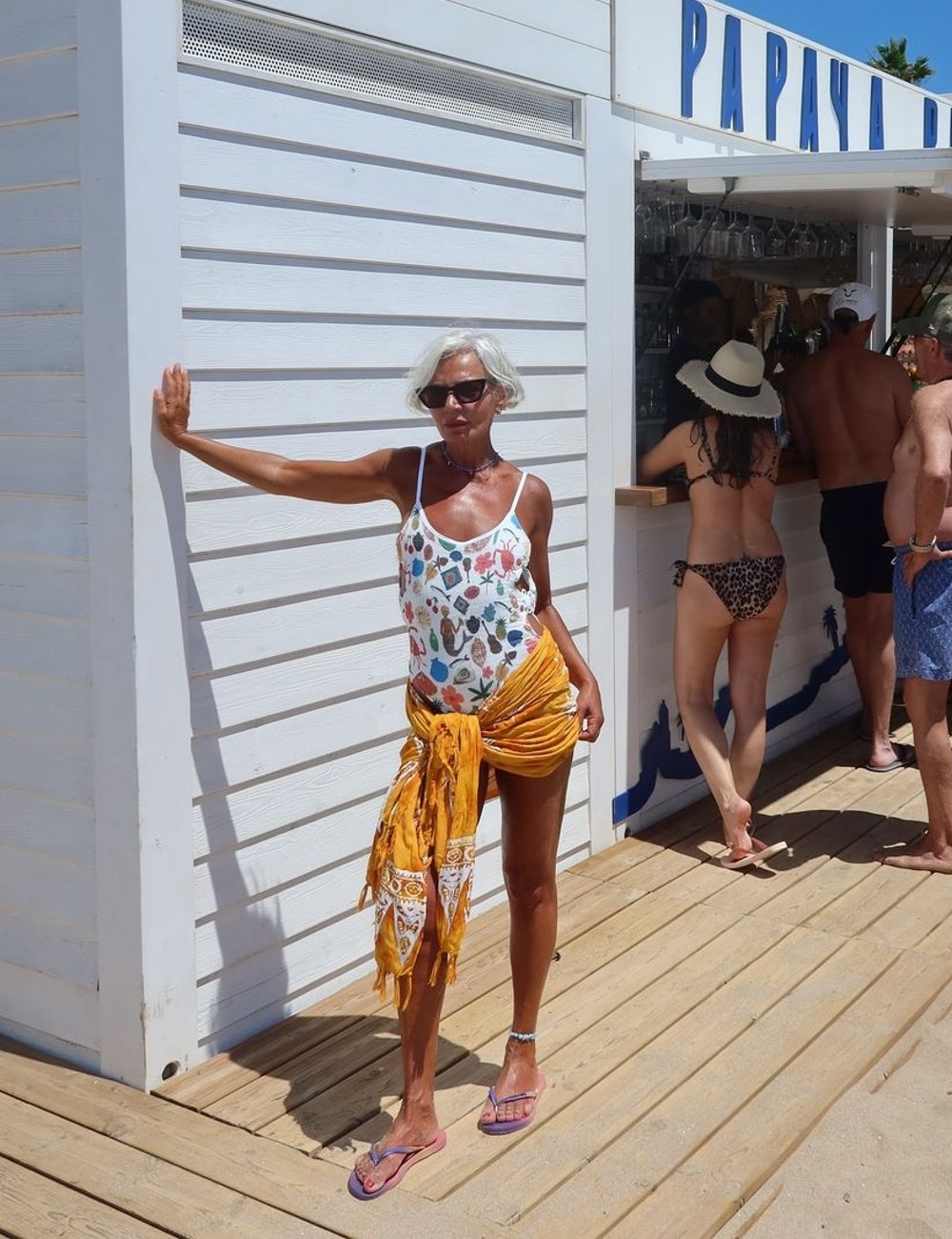 Mulher com cabelo curto e grisalho usa maiô branco estampado com frutas e saída de praia amarela amarrada na cintura. Completa o look com sandálias de dedo rosa e óculos de sol preto, destacando um estilo praia casual e colorido.