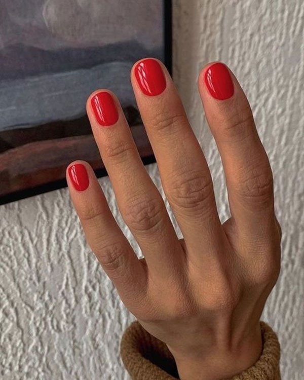 Unhas curtas e bem cuidadas com esmalte vermelho vibrante, tendência de moda minimalista e elegante. Fundo cinza e marrom claro, mangas de suéter marrom claro sugerem estilo casual e aconchegante.