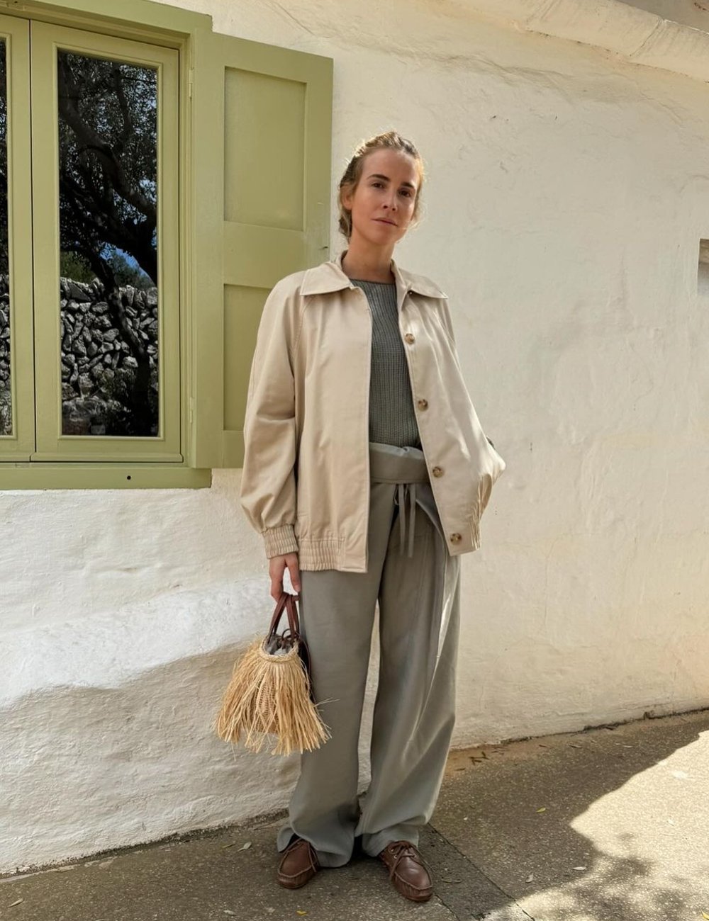 Pessoa vestida com estilo casual chic, usando jaqueta bege oversized, pulôver cinza, calça soltinha e sapato dockside. A bolsa é de palha com alça curta, conferindo um toque rústico ao look. Foco em tons neutros e tecidos confortáveis.