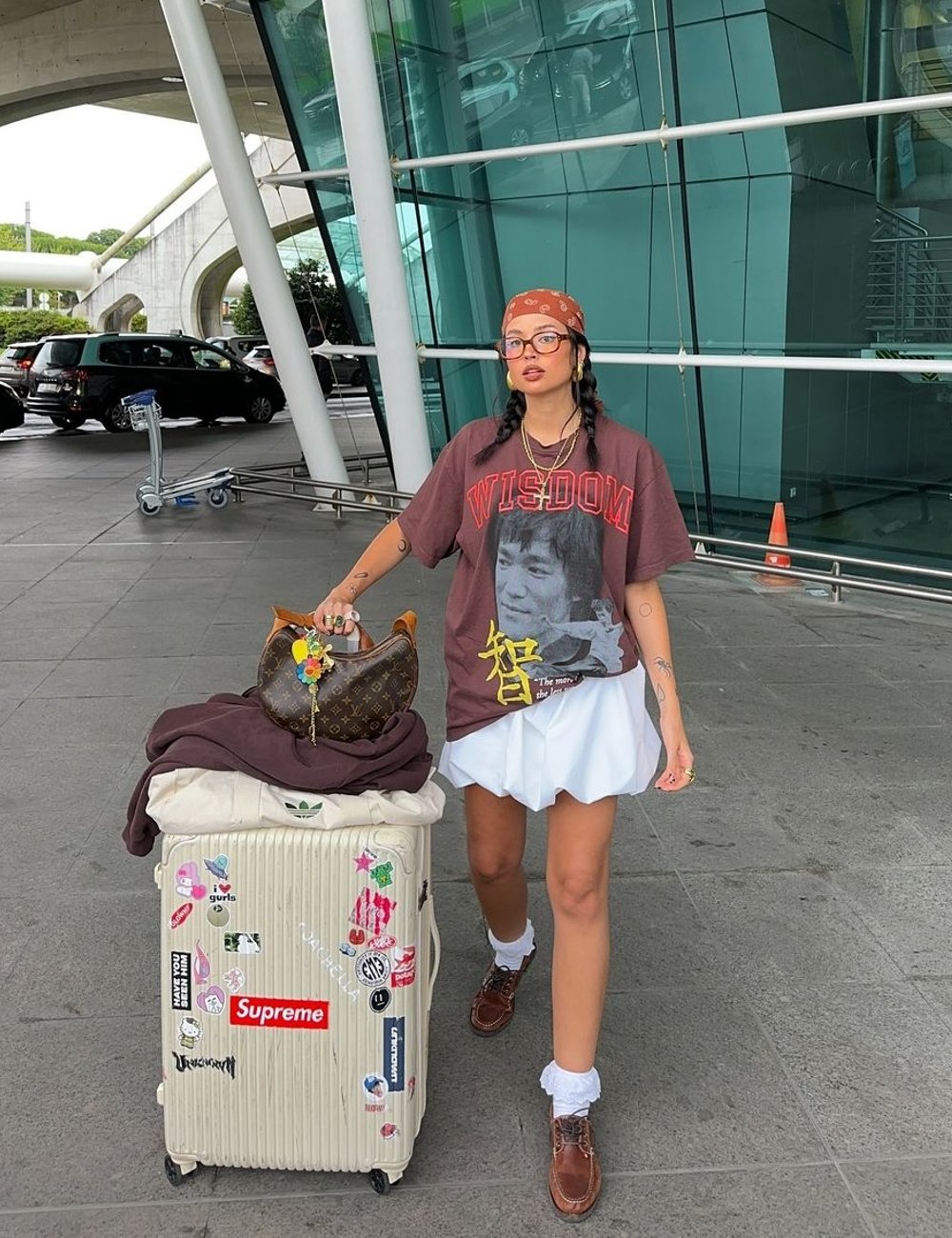 Mulher com estilo casual e urbano em aeroporto, usando camiseta oversized marrom, bandana laranja, saia branca curta, meias brancas e sapato dockside. Óculos grandes e tranças completam o look.