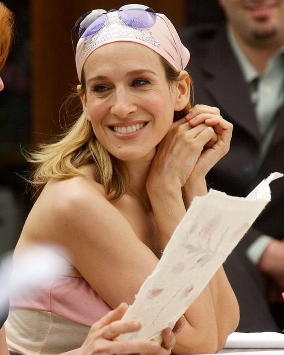 Mulher loira sorrindo, usando vestido tomara que caia e lenço rosa na cabeça, estilo bandana. Óculos de sol sobre o lenço acrescentam um toque descontraído.