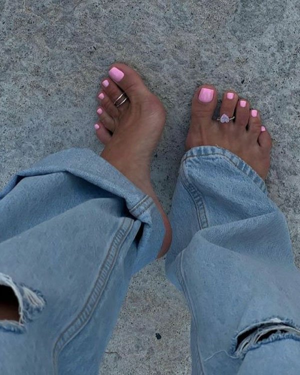 Imagem de pés com unhas pintadas de rosa claro, usando anéis nos dedos dos pés. Vestindo calça jeans larga, estilo oversized, que cobre parcialmente os pés.