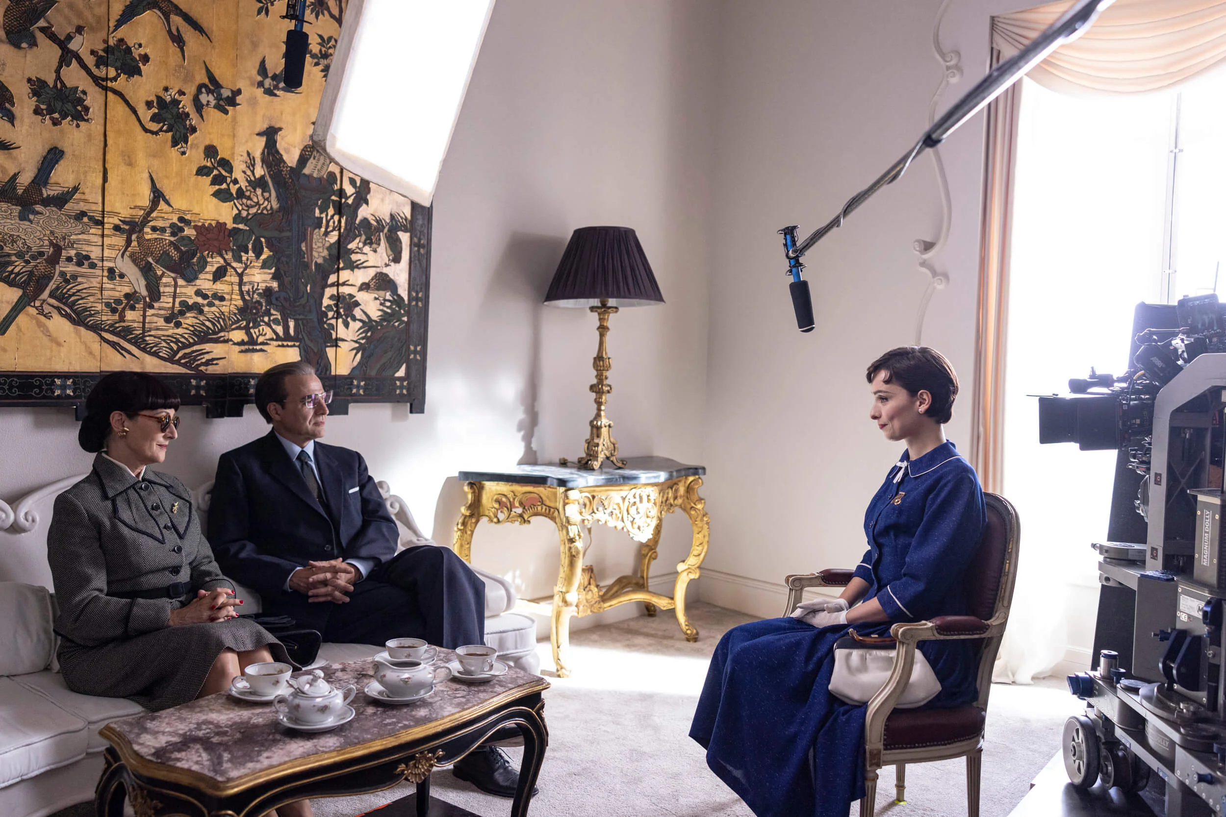 gravação, três pessoas em ambiente elegante. Casal veste trajes formais: homem de terno escuro e gravata, mulher de vestido cinza, óculos escuros e broche. Terceira pessoa em vestido azul clássico com detalhes brancos. séries biográficas