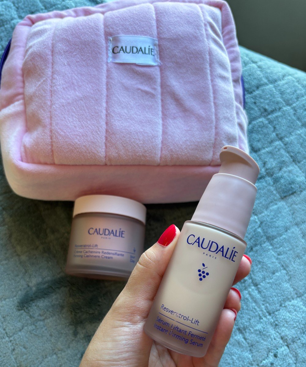 Produtos de skincare Caudalie, incluindo sérum e creme firmador Resveratrol-Lift, em destaque sobre uma almofada rosa e macia. Trends de beleza de luxo e autocuidado com foco em cuidados antienvelhecimento. Fundo com textura verde suave. sérum facial