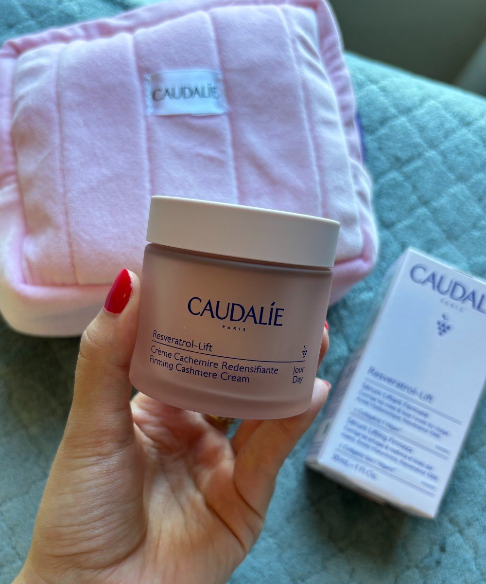 Imagem de produtos Caudalie, incluindo um creme firmador de cashmere e um acessório rosa acolchoado da mesma marca ao fundo. A mão com unhas pintadas de vermelho segura um pote do creme, destacando cuidados com a pele e bem-estar. sérum facial