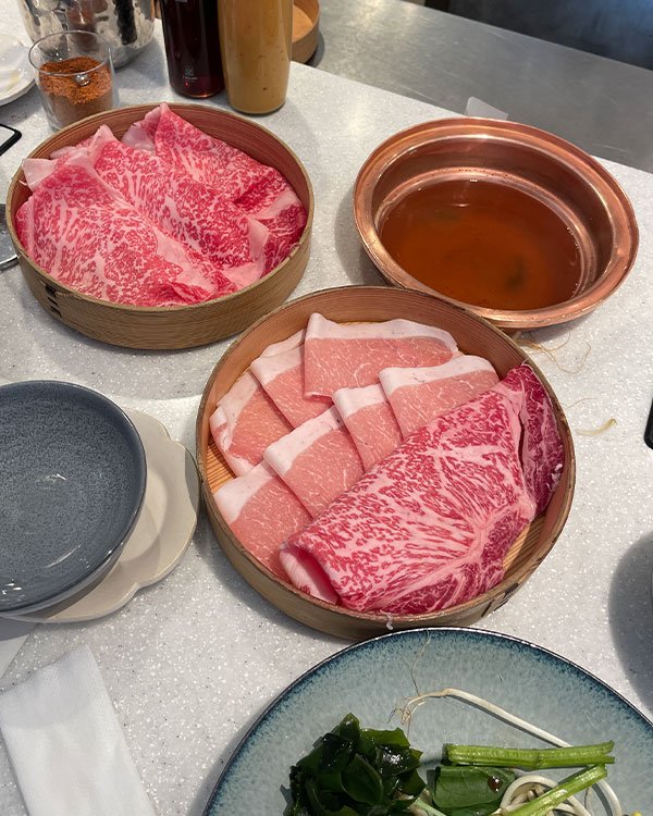 Imagem de uma refeição tipicamente japonesa com foco em pratos de shabu-shabu. Cortes finos de carne Wagyu e porco estão dispostos em cestas de bambu ao lado de um recipiente de cobre com caldo.