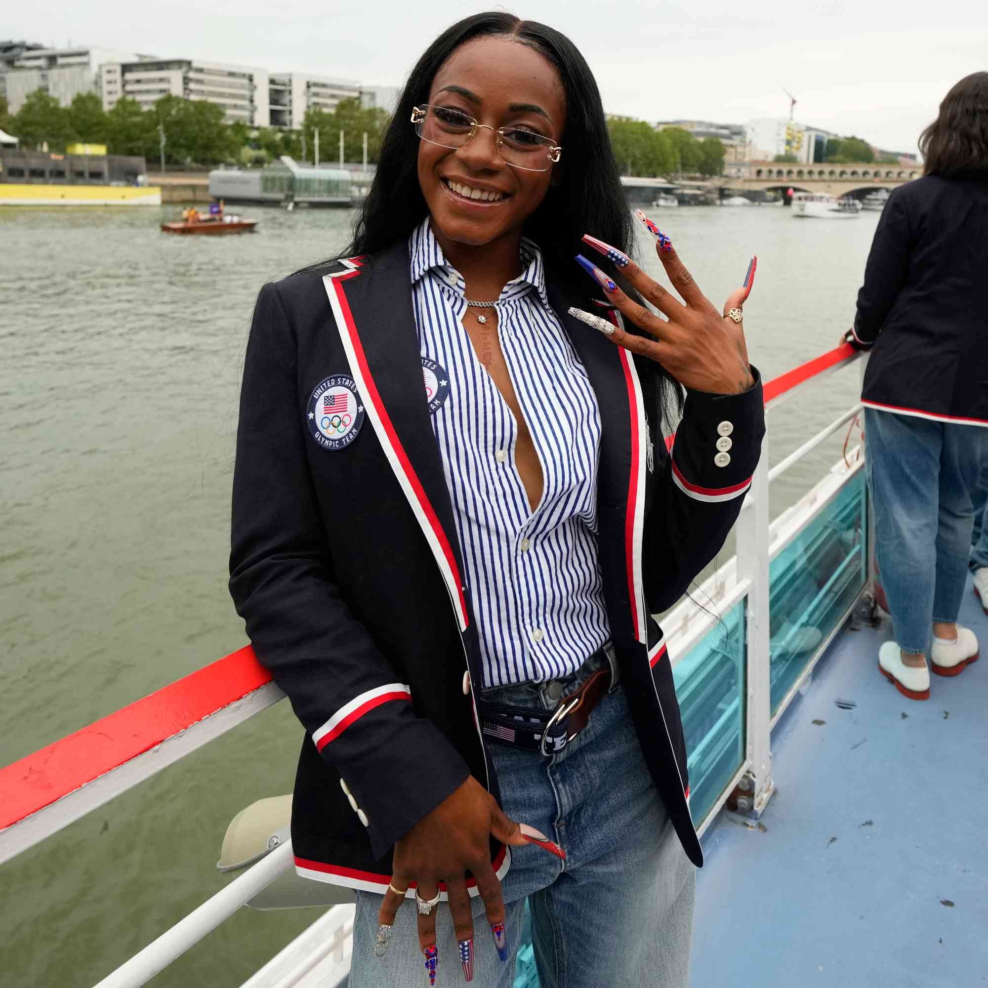 vestida com blazer esportivo azul marinho com detalhes em vermelho e branco, camisa listrada azul e branca, jeans e cinto de couro. Usa óculos de aro fina, exibe unhas longas e decoradas. Tendências esportivas e náuticas em destaque. olimpíadas 2024