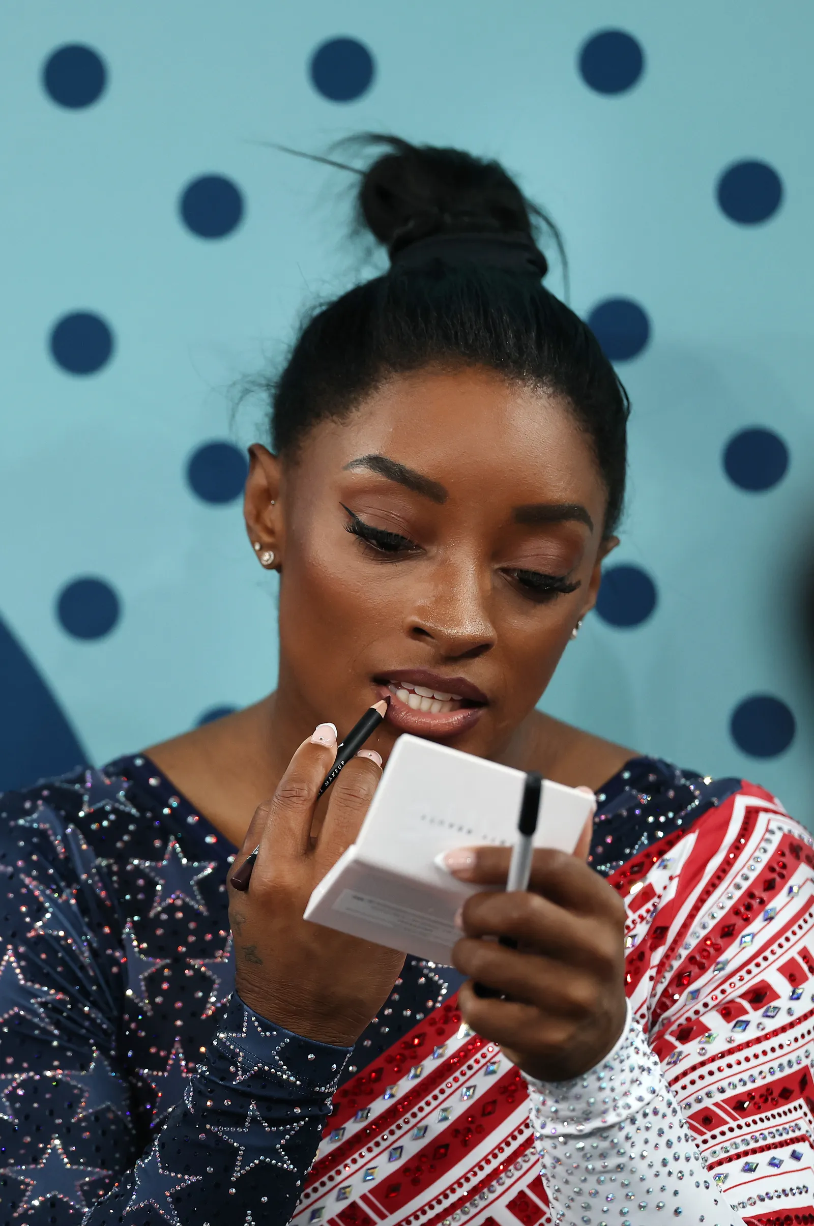 Mulher se maquiando com lápis labial, usando um maiô de ginástica brilhoso nas cores azul e vermelho, decorado com estrelas e lantejoulas. O cabelo está preso em um coque alto, refletindo uma tendência de beleza esportiva e funcional. olimpiadas 2024