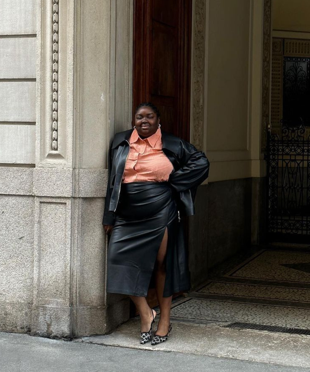 Mulher usando moda urbana chic com jaqueta e saia midi de couro preto com fenda lateral, camisa social laranja e sapatos de salto estampados. Estilo moderno e confiante destacando tendências de couro e combinações ousadas de cores e texturas.